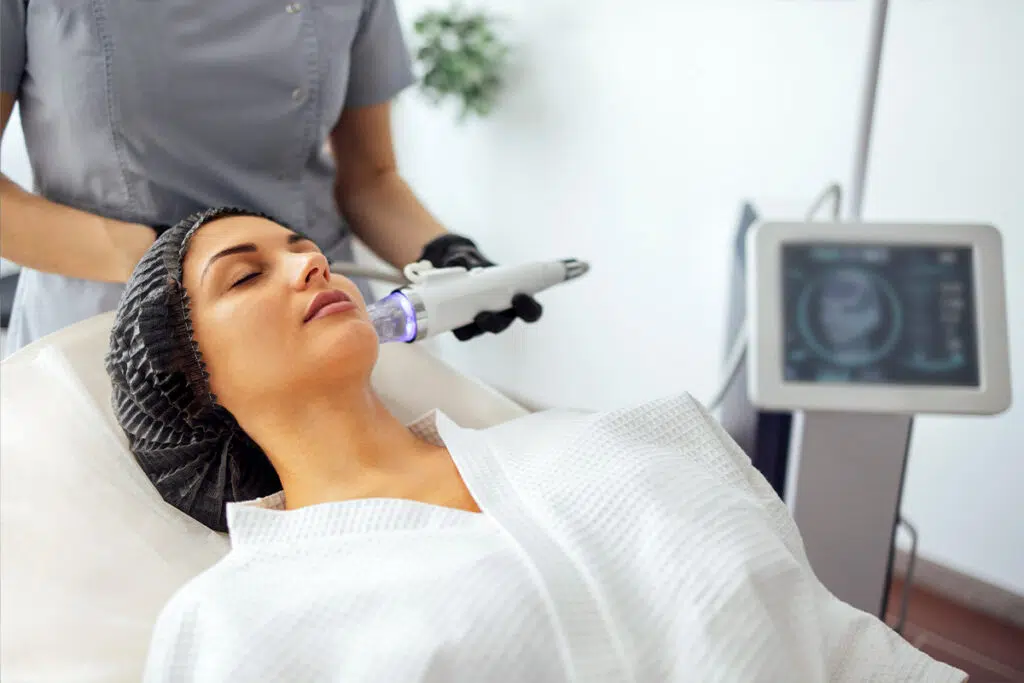 Woman reclining in a treatment chair while a clinician passes a radiofrequency handpiece over her cheek, a device monitor beside them